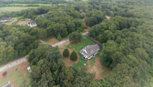 Aerial view of a large house surrounded by dense green trees, open grassy areas, and a few nearby houses scattered along winding roads in a rural landscape.