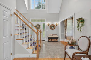 A bright entryway with a wooden staircase, tiled floor, and light gray walls. There’s a bench with storage baskets, a round mirror, a decorative wreath, and a window above letting in natural light.