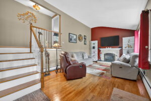 A cozy living room with hardwood floors, a staircase, two recliners, a sofa, a brick fireplace with a TV above it, white shelves, wall art, and red accent wall with matching curtains. Natural light fills the space.