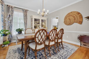 Elegant dining room with a wooden table, six cushioned chairs, a patterned rug, chandelier, white display cabinet, plants by large windows, and a woven wall art piece on a light gray wall.