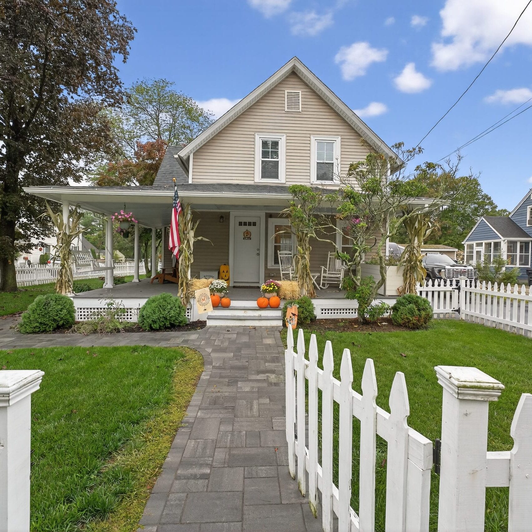 A beige two-story house with a wraparound porch, white picket fence, and landscaped yard. Pumpkins and fall decorations accent the front door. Book real estate photography CT to capture every charming detail of this inviting home.