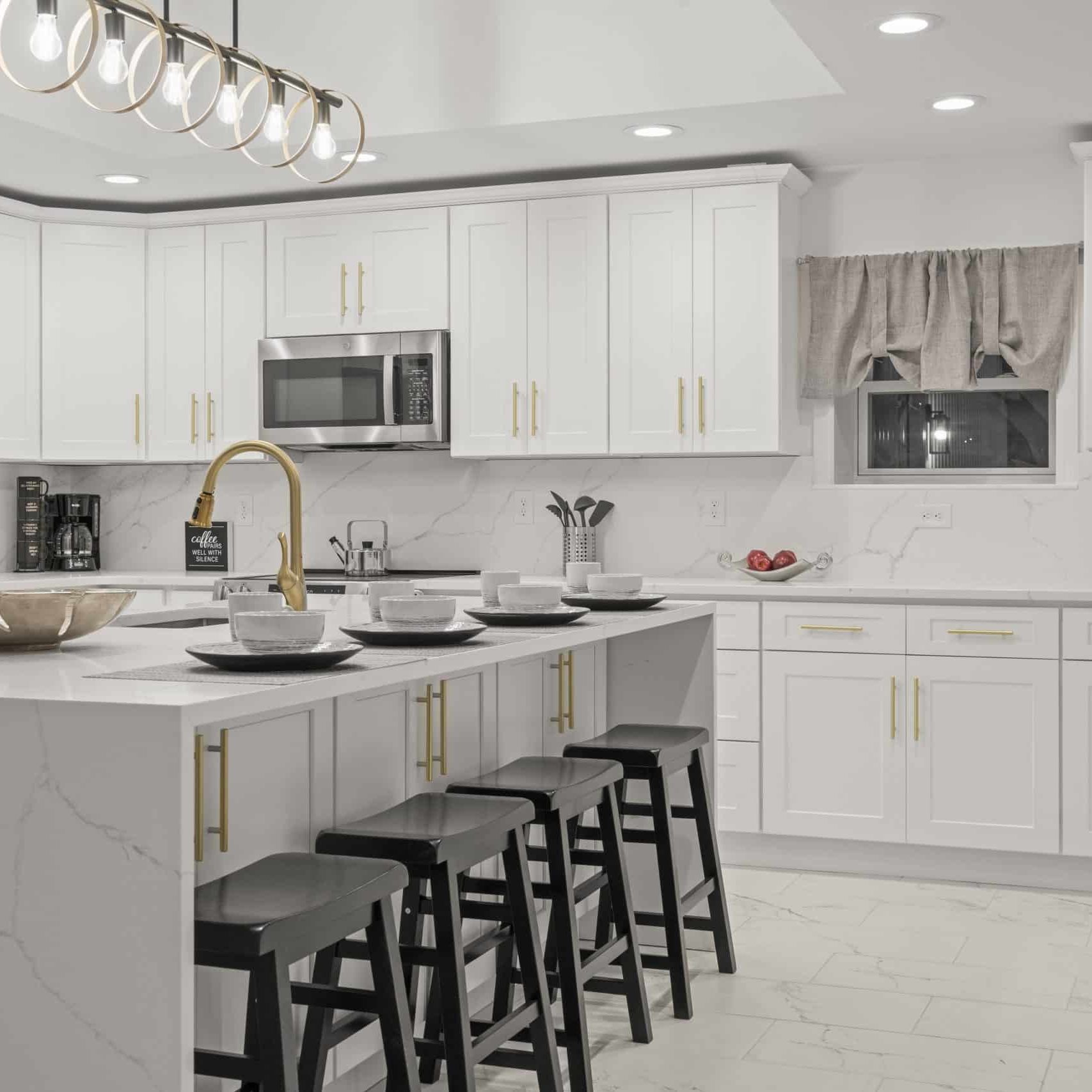 45 Westridge Rd - 74 Modern kitchen with white cabinets and countertops, gold hardware, real estate media–inspired styling, a gold faucet, stainless steel appliances, place settings on the island, four black stools, and recessed lighting.
