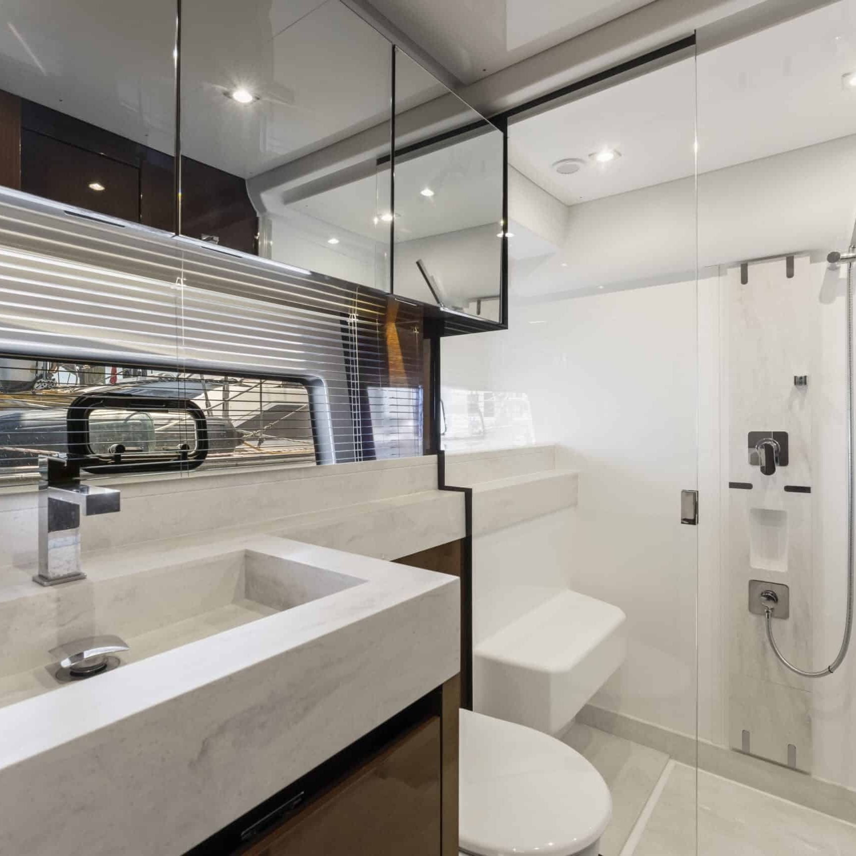 Modern bathroom with white marble sink and countertop, wall-mounted faucet, mirror cabinets, glass shower enclosure with rainfall showerhead, toilet, and large window with horizontal blinds.